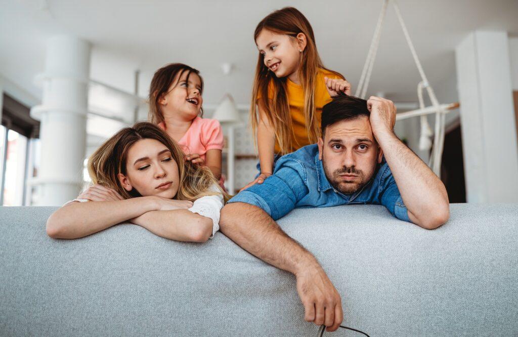 Erschöpfte Eltern auf Sofa, Kinder spielen laut dahinter | Familienservice